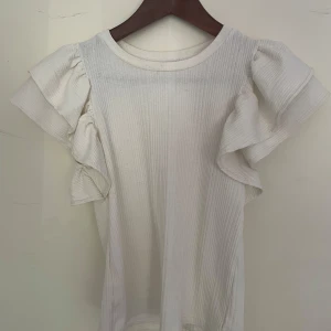 Vit ribbad topp med volangärmar Zara - Supersöt vit ribbad topp från Zara med korta volangärmar och rund halsringning. Toppen är croppad och har en mjuk, stretchig känsla som gör den riktigt skön🩷 Lite genomskinlig men inte mycket💕