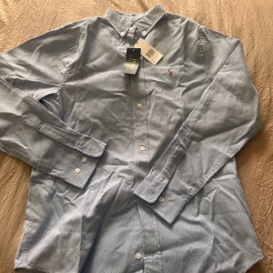 Ljusblå skjorta från Polo Ralph Lauren - Klassisk ljusblå skjorta från Polo Ralph Lauren med button down-krage och broderad logga på bröstet. Skjortan har långa ärmar, vita knappar och är tillverkad i mjuk bomull. Perfekt för en clean och stilren look.