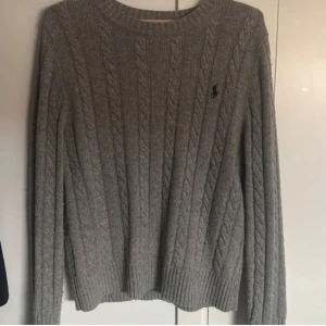 Grå kabelstickad tröja från Ralph Lauren - Kabelstickad grå tröja från Ralph Lauren med klassisk rund halsringning och diskret broderad logga på bröstet. Tröjan har långa ärmar och ribbade muddar för en snygg och tidlös look. Perfekt för lager-på-lager under kyliga dagar.