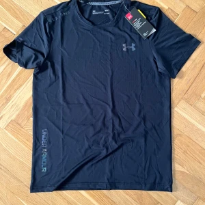 Under Armour T-shirt - Svart t-shirt från Under Armour med diskret logga på bröstet och tryck längs sidan. Tillverkad i ett lätt och snabbtorkande material som passar perfekt för träning. Klassisk rund hals och korta ärmar för en sportig look.