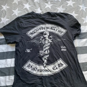 Svart Mötley Crüe t-shirt - Cool svart t-shirt med stort Mötley Crüe-tryck på framsidan och texten 'Hollywood, CA' samt 'Est. 1981'. Trycket har en fet orm med vingar och dödskalle. Klassisk passform och rund hals, perfekt för dig som gillar rock och bandmerch.