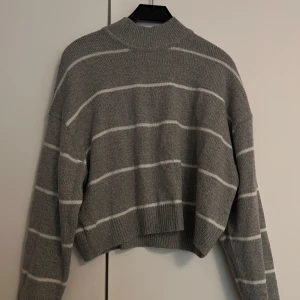 Grå randig stickad tröja - Grå stickad tröja med vita ränder och högre krage. Tröjan är croppad och har långa ärmar. Använd ett fåtal gånger.