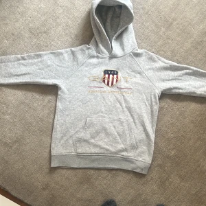 Grå hoodie från GANT med broderi - Säljer en grå hoodie från GANT med broderat emblem och texten 'American Sportswear' på bröstet. Tröjan har huva, magficka och långa ärmar. Materialet är mjuk bomull och passformen är normal. Perfekt för chill dagar och sportig stil. En snygg tröja för varje dag. Den funkar till en grish outfit men även till chill hemma. Storleken är får 13 till 14 år!