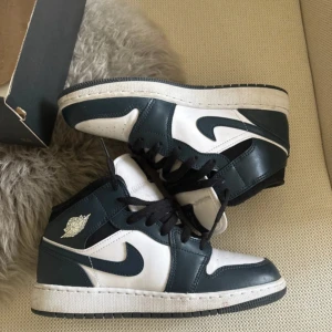 Nike Air Jordan 1 Mid svart/vit - Snygga Nike Air Jordan 1 Mid sneakers i svart och vitt skinn med klassisk siluett. Skorna har svarta snören, perforerad tå och ikoniska Jordan Wings-loggan på sidan. Perfekta för dig som vill ha en clean och tidlös look.