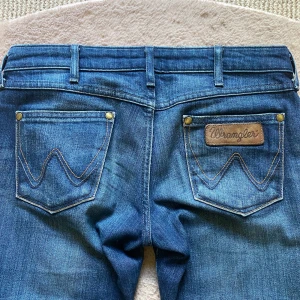 Lågmidjade utsvängda jeans - Snygga blå bootcut low waist jeans från vintage Wrangler med klassiska detaljer och logga på bakfickan. Jeansen har normal passform, fem fickor och är tillverkade i slitstarkt denim. Perfekta för dig som gillar retrovibbar och vill ha ett par jeans med lite utsvängda ben!😻Passar till varje plagg💋Innerben 80cm, midja 36cm, grenhöjd 19cm! Storlek W26 & L32, passar XS💕Billigare på min Vinted: 4annie1. Pris kan diskuteras❤️