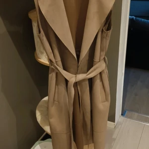 Beige lång väst från Zara med bälte - Snygg beige lång väst från Zara med breda slag och knytskärp i midjan. Västen har djupa fickor och är tillverkad i ett mjukt material som påminner om mocka. Perfekt för lager-på-lager och ger en clean look.