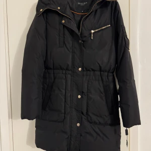 Svart dunjacka från Hollies utan päls  - Svart lång dunjacka från Hollies med guldiga dragkedjor och knappar. Jackan har huva, flera fickor och en snygg detalj med logotyp på ärmen. Perfekt för kalla dagar och har en klassisk, stilren look.