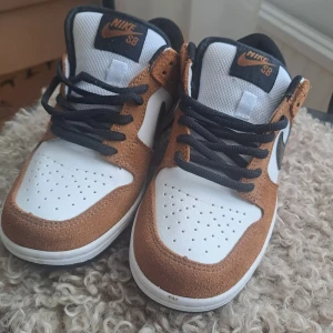 Nike SB Dunk Low i brunt och vitt - Nike SB Dunk Low sneakers med vit läderbas, bruna mockadetaljer och svart Swoosh i struktur. Skorna har svarta skosnören, svart yttersula och mesh på plösen. Perfekt för dig som gillar streetwear och skatekultur. Skon tillhör en samlarserie och är tillverkad I begränsad upplaga. Strl.36