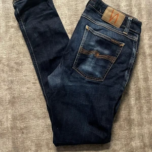 Mörkblå jeans från Nudie Jeans - Snygga mörkblå jeans från Nudie Jeans med klassiska kontrastsömmar och ikonisk bakficka. Jeansen har en smal passform och är tillverkade i mjukt denimtyg. Perfekta för dig som gillar stilrena och tidlösa jeans med en modern touch.