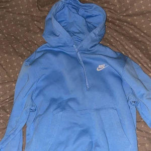 Blå Nike hoodie med huva - Snygg blå hoodie från Nike med klassisk logga broderad i vitt på bröstet. Tröjan har huva med snörning och en stor magficka framtill. Perfekt för dig som gillar sportig och avslappnad stil.