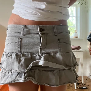 Grå volangkjol från Minga London - Supersnygg grå miniskirt från Minga London med jeanslook och volanger. Kjolen har fickor fram, bälteshällor och en cool volangkant som ger extra attityd. Perfekt för dig som gillar streetstyle och vill sticka ut med en edgy vibe.