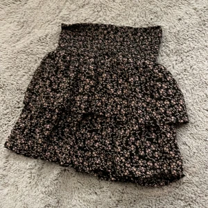 Svart blommig kortkjol med volang - Supersöt svart kortkjol med ljusrosa blommigt mönster och bred smock i midjan. 