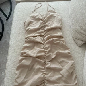 Beige omlott kortklänning med rynk - Säljer en beige kortklänning med tunna axelband och snygg rynkdetalj längs hela framsidan. Klänningen har omlott-look och är perfekt för sommarens alla tillfällen. Materialet känns lätt och luftigt, och färgen är neutral och enkel att matcha.