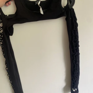 Svart långärmad topp med snörning - Cool svart långärmad topp från Bershka med snörningsdetaljer längs hela ärmarna som ger en edgy och unik look. Toppen är figurnära och har en öppen design på ärmarna med flätad snörning. Perfekt för dig som vill sticka ut.