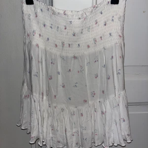 Vit blommig kortkjol med volang - Supersöt vit kortkjol med små blommor i rosa och blått. Kjolen har smock upptill och en bred volang längst ner som ger en flowy känsla. Perfekt för dig som gillar romantisk och feminin stil. Materialet känns lätt och luftigt.