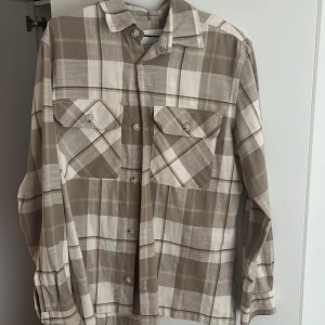 Rutig overshirt från Jack & Jones - Snygg rutig overshirt från Jack & Jones i beige och grå toner. Skjortan har två stora bröstfickor med lock och knappar framtill. Perfekt att bära öppen över en t-shirt eller stängd som en vanlig skjorta. Materialet känns mjukt och bekvämt. Nypris 450 säljer för 120