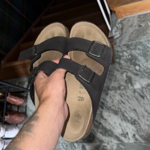 Svarta sandaler från Birkenstock - Klassiska svarta sandaler från Birkenstock med två breda remmar och justerbara metallspännen. Fotbädden är i kork och mocka för extra komfort. Perfekta för sommaren och riktigt sköna att glida runt i.