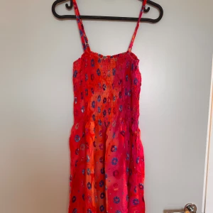Röd/orange sommarklänning - Färgglad röd/orange klänning med ett mönster av blå cirklar. Den har smala axelband och en smockad överdel som ger en bekväm passform. Klänningen är lätt och luftig, vilket gör den bekväm att bära. Perfekt för både stranddagar och sommarkvällar. 