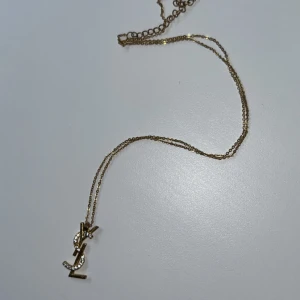 Guldhalsband med YSL-berlock - Elegant guldhalsband med en YSL-berlock dekorerad med små glittrande stenar. Perfekt för att ge en lyxig touch till din outfit. Kedjan är tunn och smidig, vilket gör den bekväm att bära hela dagen.