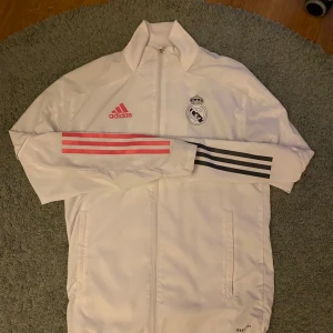Vit träningsjacka från Adidas - Säljer en vit träningsjacka från Adidas med Real Madrid-logga. Jackan har dragkedja och långa ärmar med rosa och svarta ränder. Perfekt för fotbollsfans! 💪