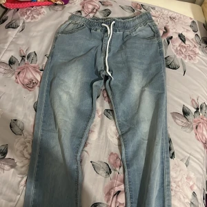 Ljusblå jeansbyxor med snörning - Säljer ett par ljusblå jeansbyxor med elastisk midja och vit snörning. Byxorna har en avslappnad passform och är perfekta för en bekväm stil. De har fickor fram och är gjorda av ett mjukt denimtyg.