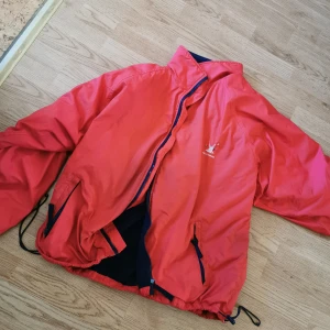 Röd vindjacka från Helly Hansen - Säljer en röd vindjacka från Helly Hansen med dragkedja och broderad logga på bröstet. Jackan har en blå fleecefoder på insidan för extra värme. Perfekt för blåsiga dagar! Väldigt varm. Gott skick. Vintage. 