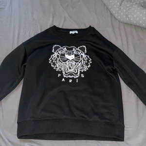 Svart tröja från Kenzo - Säljer en svart tröja från Kenzo med ett stort tigertryck och texten 'Kenzo Paris' på framsidan. Tröjan har en rund halsringning och långa ärmar. Perfekt för en stilren look.