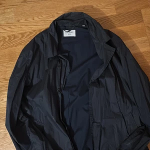 Svart kappa från Our Legacy - Snygg svart kappa från Our Legacy, tillverkad i 100% bomull med tyg från Italien. Perfekt för en stilren look. Kappan har en klassisk design med knappar framtill.