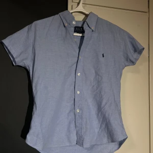 Blå kortärmad skjorta från Ralph Lauren - Säljer en stilren blå kortärmad skjorta från Ralph Lauren. Skjortan är tillverkad i 100% bomull och har en klassisk krage med knappar. Perfekt för en avslappnad men ändå elegant look (ganska liten small). 