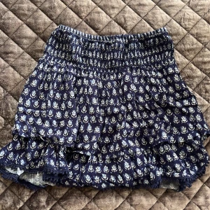 Blå mönstrad kjol från Pull&Bear - Säljer en söt blå kjol från Pull&Bear med ett vitt blommönster. Kjolen har en volangdetalj och elastisk midja för en bekväm passform. Perfekt för varma dagar!