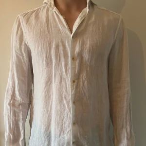 Vit linneskjorta - Säljer en stilren vit linneskjorta med lång ärm och klassisk krage. Skjortan har knappar framtill och är perfekt för en avslappnad men ändå elegant look. Passar bra till både jeans och chinos.