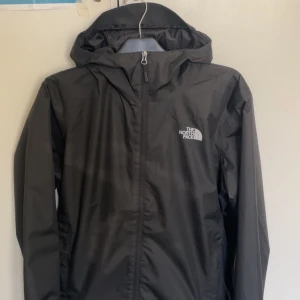 Svart vindjacka från The North Face - Säljer en svart vindjacka från The North Face med huva och dragkedja. Perfekt för blåsiga dagar och har en stilren design med logotyp på bröstet och ryggen.