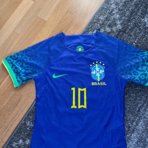 Blå Brasilien fotbollströja med Neymar JR - Säljer en blå Brasilien fotbollströja från Nike med Neymar JR och nummer 10 på ryggen. Tröjan har kortärmade ärmar med ett leopardmönster i grönt och blått. Perfekt för fotbollsfans!
