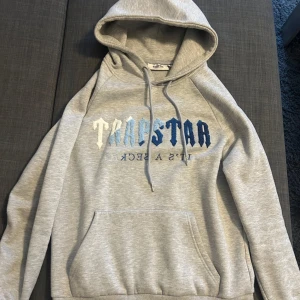 Grå hoodie från Trapstar - Säljer en snygg grå hoodie från Trapstar med tryck i blå och vit på bröstet. Den har en klassisk känguruficka och justerbar huva med dragsko. Perfekt för en casual look eller att mysa i hemma. Passar till alla årstider! Storlek S.