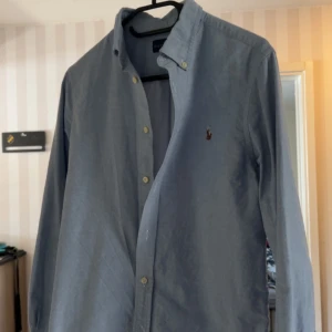 Blå skjorta från Ralph Lauren - Snygg blå skjorta från Ralph Lauren med klassisk krage och knappar framtill. Skjortan har en liten broderad logotyp på bröstet och är långärmad. Perfekt för en stilren look. Skick 9/10 storlek 16 år/xs