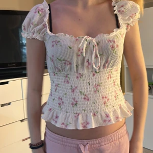 Vit blommig smocktopp med volangdetaljer - Säljer en söt vit smocktopp med rosa blommor och gröna blad. Toppen har korta puffärmar och volangdetaljer vid ärmslut och nederkant. Perfekt för en somrig look! 🌸