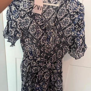 Mönstrad klänning från ZARA - Snygg mönstrad klänning från ZARA i blått och vitt. Klänningen har en v-ringad design och korta ärmar, perfekt för en stilfull look. Den har ett unikt mönster som ger en trendig touch.
