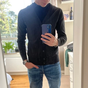 Svart kofta med knappar - Säljer en stilren svart kofta med knappar framtill. Den har en v-ringad design och långa ärmar, perfekt för en elegant look. Passar bra till jeans eller chinos.