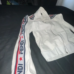 Vit jacka från Fendi - Säljer en snygg vit jacka från Fendi med dragkedja och ribbade muddar. Jackan har långa ärmar med Fendi-logga och röda stjärnor längs sidorna. På baksidan finns en stor Fendi Roma-brodering i blått och rött. Perfekt för dig som vill ha en stilren och exklusiv look. storleken är L, men sitter som M.