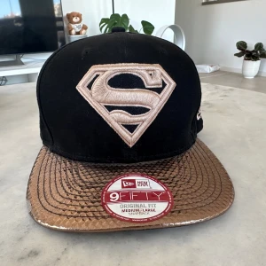 Svart och guld Superman keps från New Era - Snygg svart keps med guldfärgad skärm och broderad Superman-logga framtill. Kepsen är av modellen 9FIFTY och har en justerbar passform. Perfekt för dig som vill sticka ut med en unik stil.liten repa på sidan.