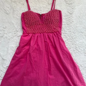 Rosa klänning med pärldetaljer - Snygg rosa klänning med pärlor på överdelen och smala axelband. Klänningen har en dragkedja bak och en v-ringad design framtill. Perfekt för en festlig look! Observera att det finns ett par lösa trådar, dock syns inte dessa på håll💗💗