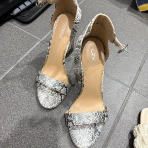 Ormskinnsmönstrade klackskor från Catwalk - Snygga klackskor med ormskinnsmönster från Catwalk. Skorna har en öppen tå och en elegant remdesign med spänne runt ankeln. Perfekta för att ge en trendig touch till din outfit.