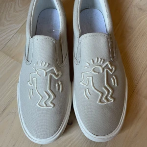 Keith Haring embroidered shoe - Snygga beige slip-on sneakers med ikonisk Keith Haring-design i relief på ovansidan. Skorna har en mönstrad sula med samma konstnärliga stil. Perfekta för dig som vill ha en unik och konstnärlig touch på dina skor. Skorna har tyvärr ingen sula, men är i övrigt i gott skick. Skicka pris är flexibelt