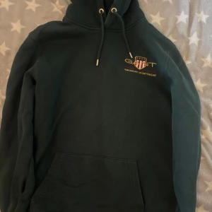  GANT hoodie - Säljer en snygg mörkgrön hoodie från GANT med broderad logga på bröstet. Tröjan har en klassisk känguruficka och justerbar huva med dragsko. Perfekt för en avslappnad stil. Inte använd mycket alls och är i bra skick