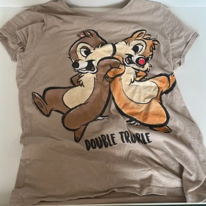 Beige Disney T-shirt  - Säljer en beige T-shirt från Disney med ett tryck av Chip och Dale och texten 'Double Trouble'. Perfekt för alla Disney-fans! T-shirten har korta ärmar och är gjord i ett mjukt material.