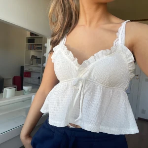 Vit volangtopp med knytband - Säljer en söt vit topp med volangdetaljer och knytband framtill. Toppen är ärmlös och har en luftig passform, perfekt för varma dagar. Aldrig använd.