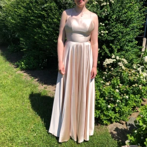 Champagnefärgad balklänning från Dressroom Linné  - Elegant champagnefärgad balklänning i satin med smala axelband och öppen rygg. Klänningen har en vacker slits framtill och knytband i ryggen för en justerbar passform. Den har även en mindre skada på höger sida vilket visas på bild 3. Fler bilder kan skickas vid intresse😊 Passar både 36-38 tack vare snörningen i ryggen. Sen är den uppsydd. Jag är 167 cm och bar ca 4 cm höga klackar då nådde klänningen precis marken. 
