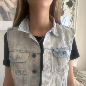 Ljusblå jeansväst med knappar - Snygg ljusblå jeansväst med knappar framtill och två bröstfickor. Perfekt för att ge din outfit en cool touch. Västen har en klassisk krage och är ärmlös.