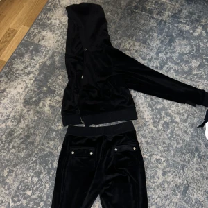 Svart velourset från Juicy Couture - Säljer ett svart velourset från Juicy Couture med dragkedja och huva. Setet har långa ärmar och byxor med snörning i midjan. Perfekt för en avslappnad och stilren look.
