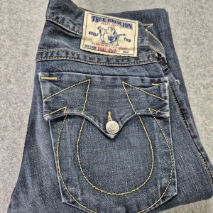 True Religion jeans med bootcut - Säljer ett par snygga True Religion jeans i mörkblå denim med bootcut-stil. De har de ikoniska bakfickorna med kontrastsömmar och en knappdetalj. Perfekta för dig som vill ha en klassisk look med en twist.